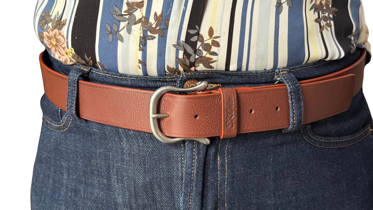 Brown Lazzaro leather belt worn with dark blue jeans and a striped floral top, showing its metal buckle, embossed leather keeper, and adjustment holes.