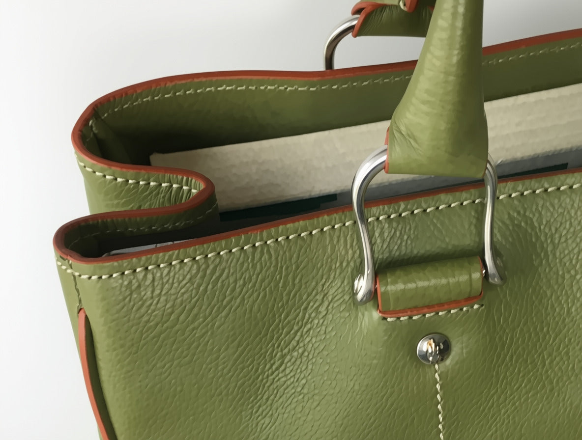 Close-up of green Allegra leather tote bag showing metal handle attachment, rivet, leather grain, contrast stitching, and reddish edge trim.