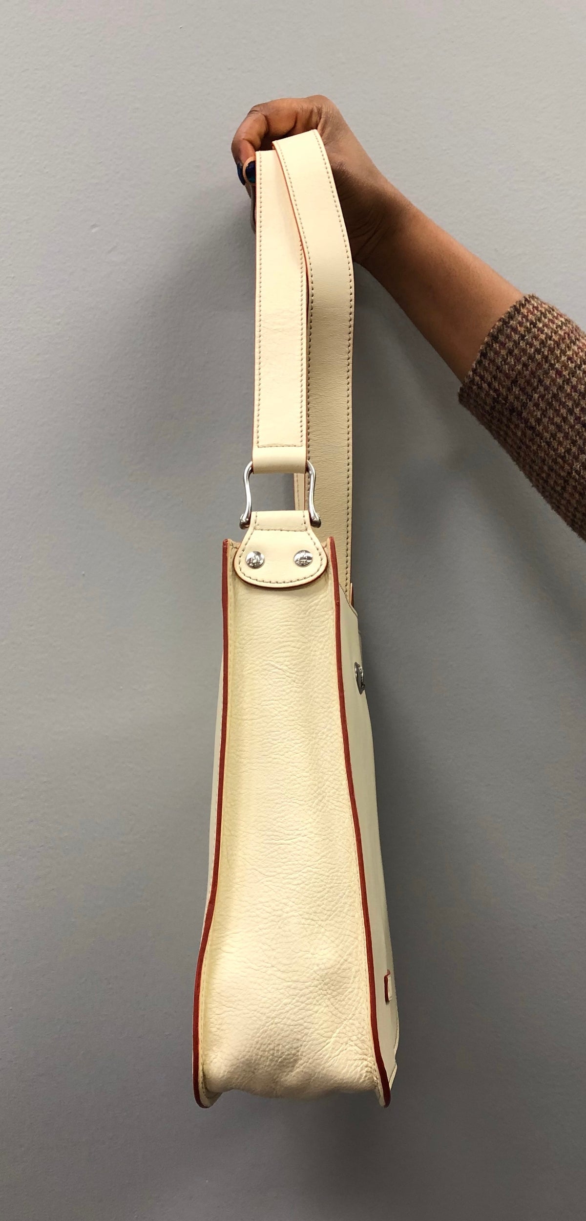 Side profile of ivory Emilia/Emilio leather messenger bag held by its strap, showing leather grain, contrasting reddish edges, and riveted metal strap attachment.