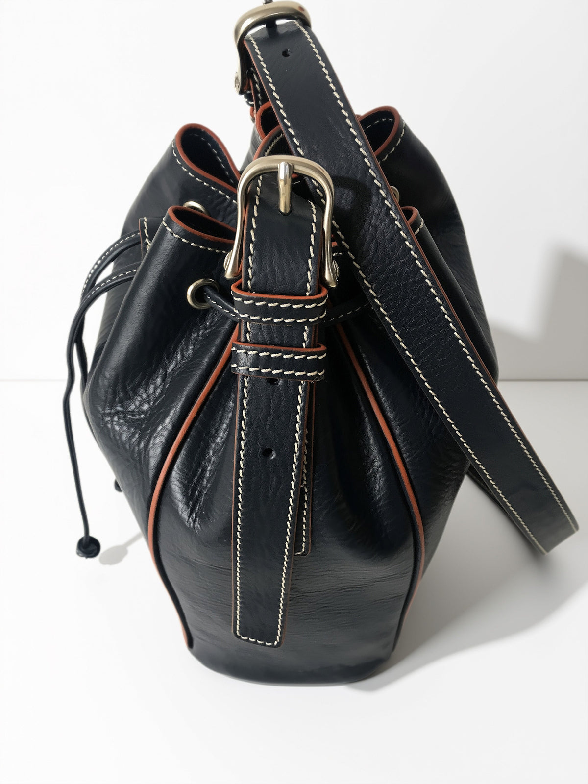 Side view of blue Febe leather bucket bag showing adjustable buckle, strap holes, keeper loops, and contrast stitching.