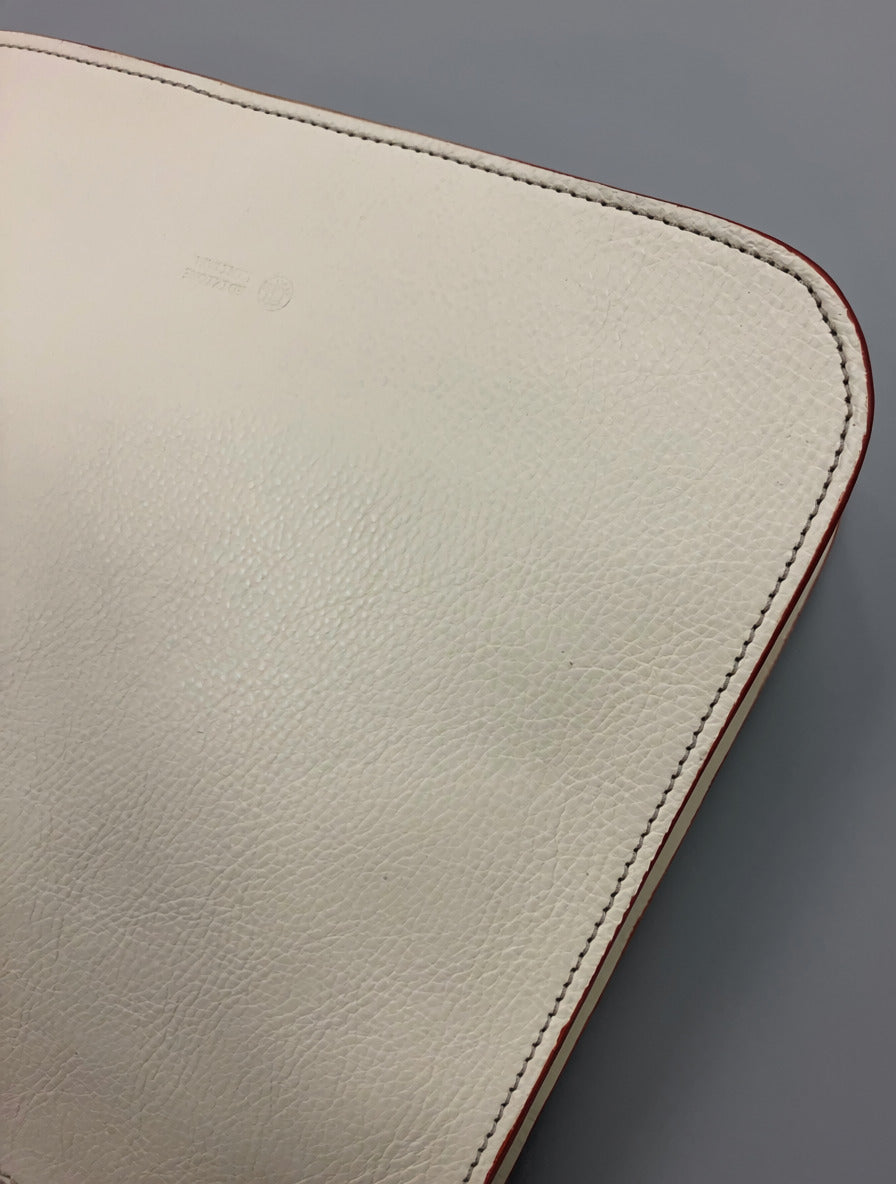 Close-up of ivory Emilia/Emilio leather messenger bag showing leather grain, stitching around a rounded corner, and contrasting reddish edge trim.